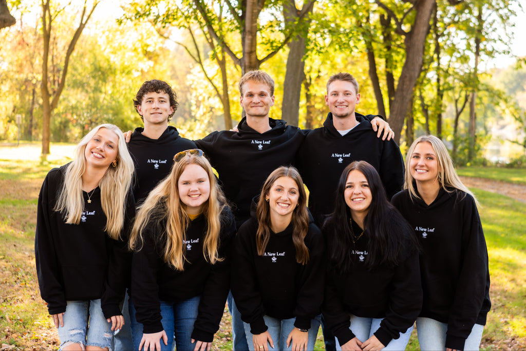 A group of nine young adults posing together outdoors in a park, wearing matching black hoodies with a white logo that reads 'A New Leaf Tea'.