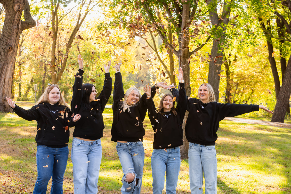 Five women standing outdoors in a park with autumn trees, wearing matching black hoodies and light-wash blue jeans, with leaves being tossed in the air.