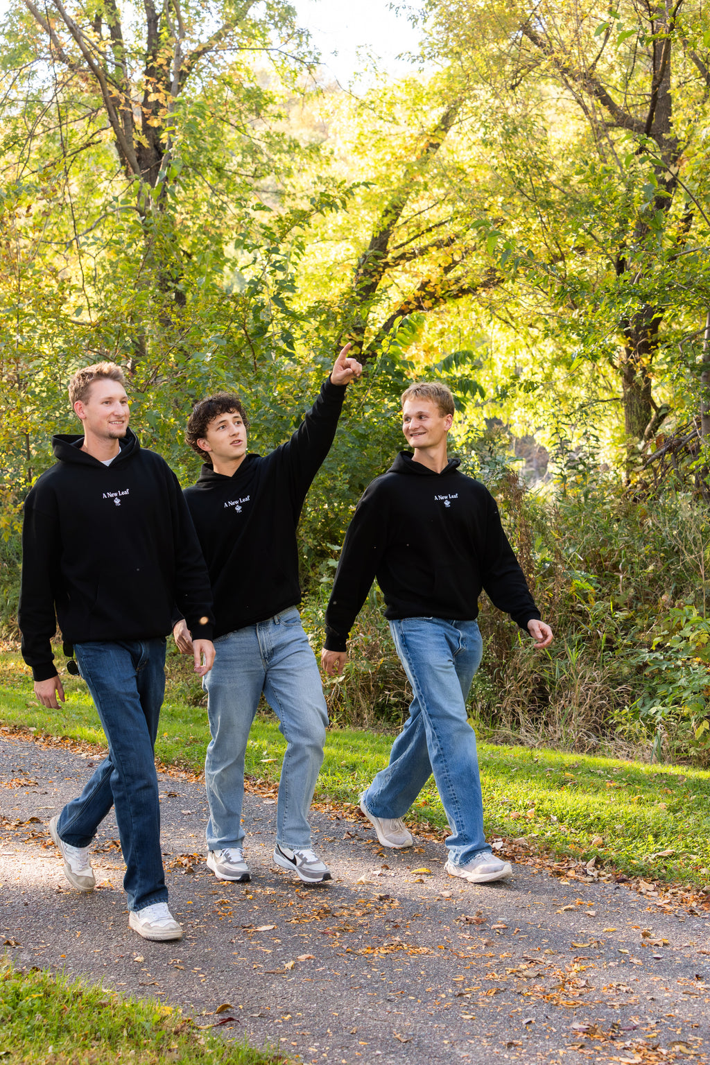 Three young men walking on a paved path in a wooded outdoor setting, wearing matching black hoodies with a small white logo on the chest, blue jeans, and white sneakers.