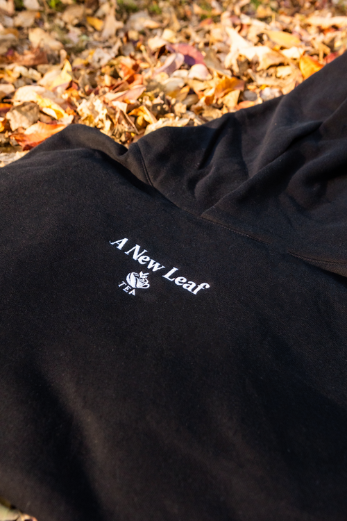 A close-up of a black hoodie laid outdoors on a bed of dry autumn leaves, featuring a white embroidered logo and the text 'A New Leaf' on the chest.