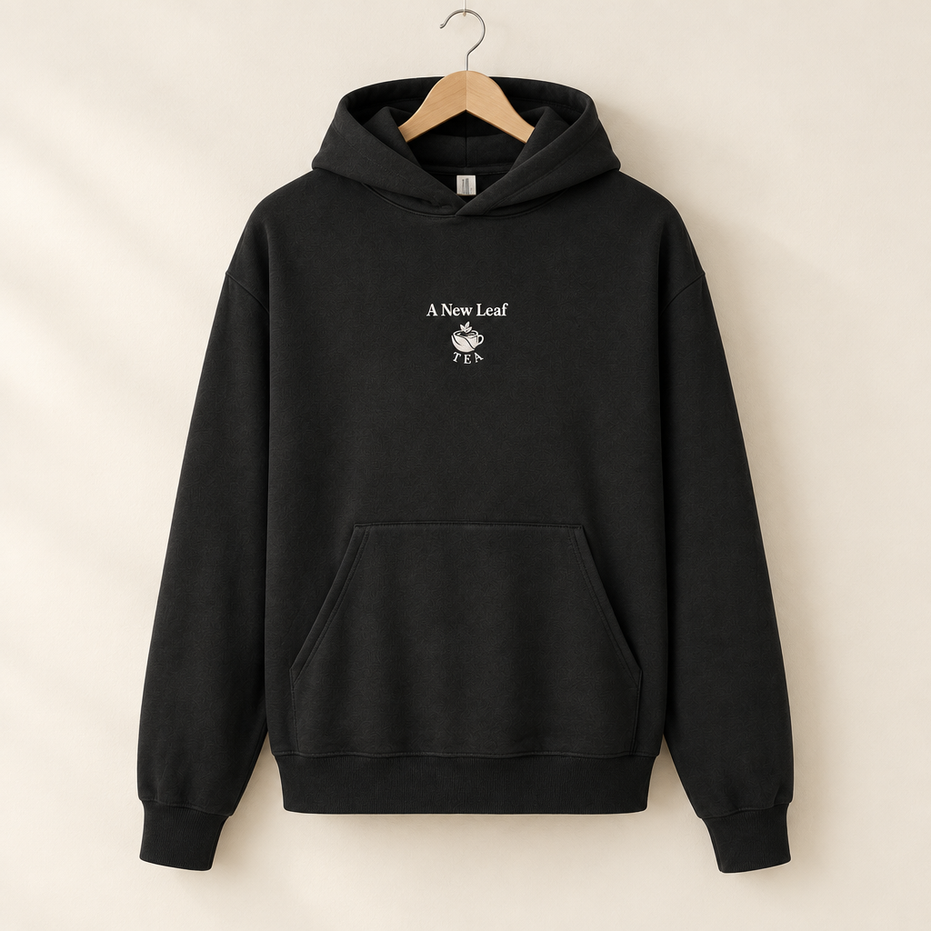A black pullover hoodie hanging on a wooden hanger against a warm beige background. The hoodie features a small white graphic on the center chest with the text 'A New Leaf' and a leaf illustration.