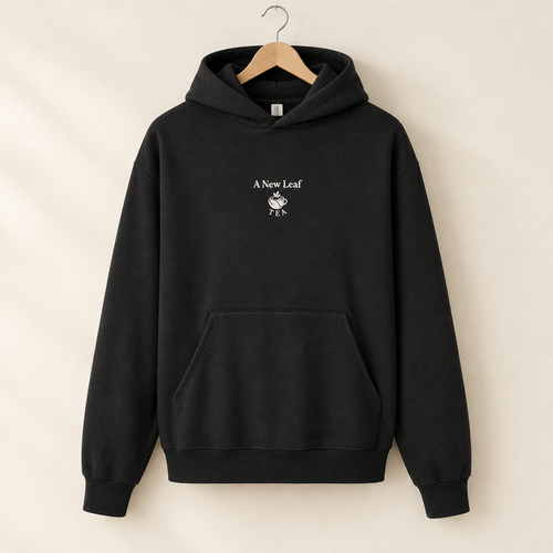 A black pullover hoodie hanging on a wooden hanger against a warm beige background. The hoodie features a small white graphic on the center chest with the text 'A New Leaf' and a leaf illustration.